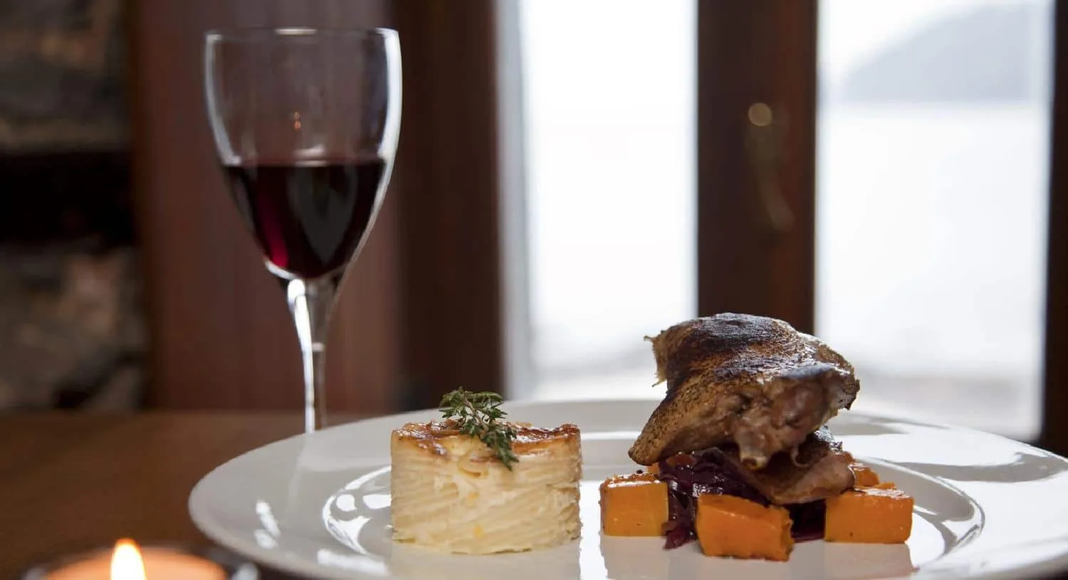 A plated duck dish with dauphinoise potatoes and roast squash, a glass of red wine behind